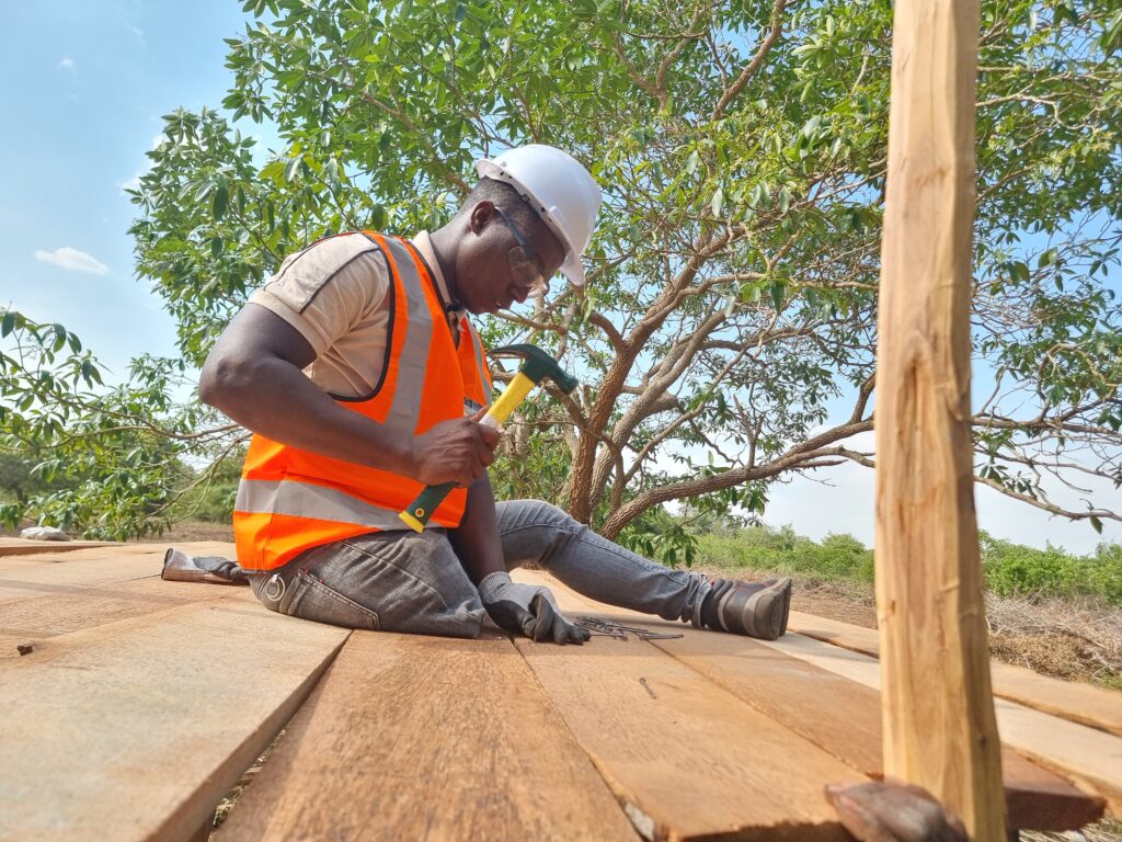 Men with physical disability dressed with a safety waistcoat, helmet and hammer sitting on wooden structure that he is currently building.