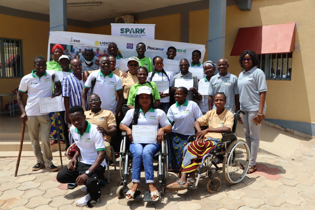 A group of women and men with and without disabilities pose in front of a banner which says SPARK on it in front of a building.