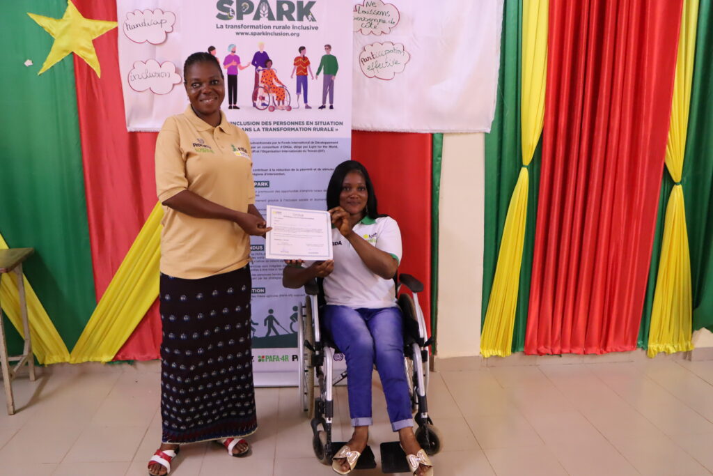 A young women in a wheelchair is given a certificate by another women in front of a SPARK banner. Both are smiling into the camera.